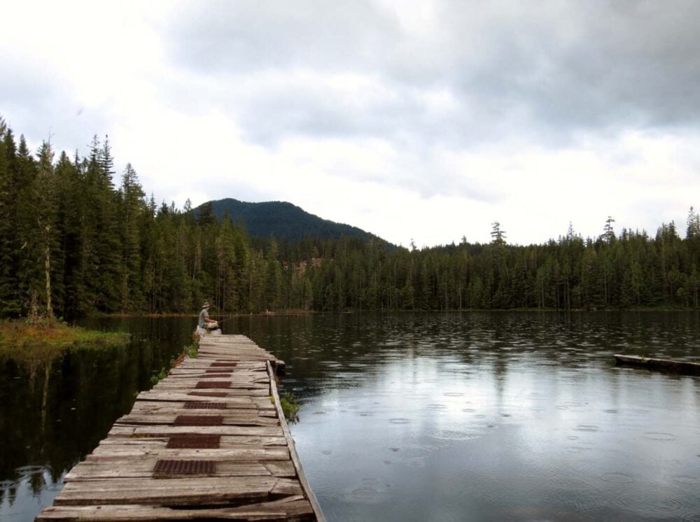 Looking down wooden lake dock to JR, who is sitting at the end and fishing in the rain. The lake is surrounded by forest