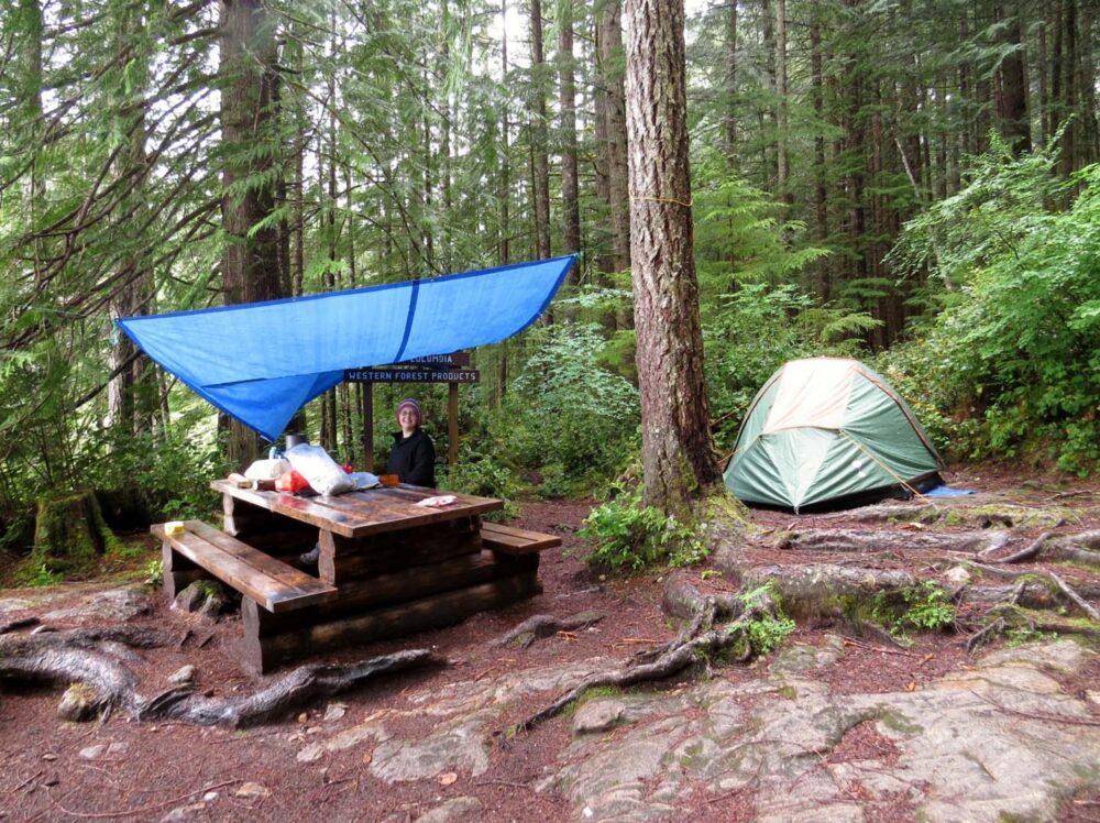Set up tent in the forest next to picnic table, with tarp set up above 
