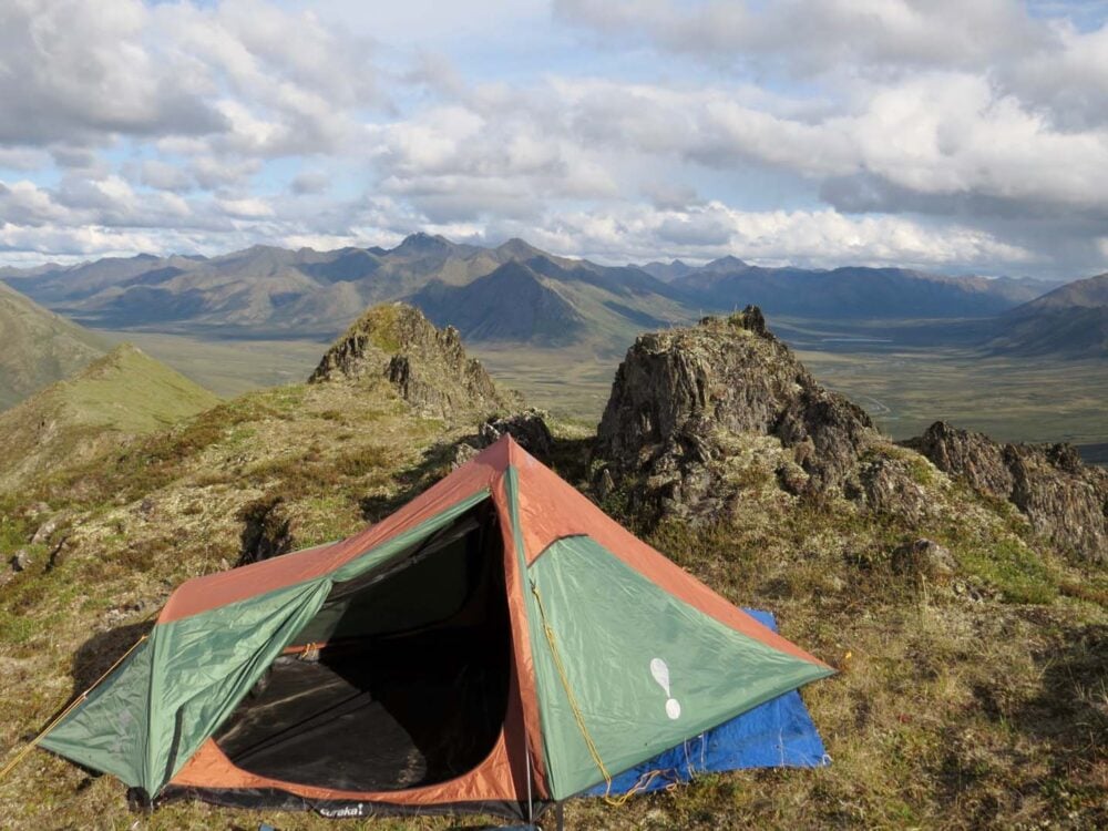 Green and orange tent pitched at top of mountain with peaks beyond