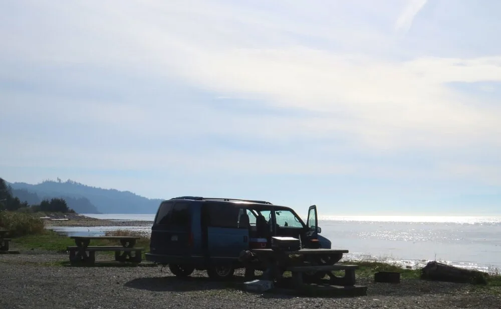Side view of blue van parked by picnic table next to calm ocean at Jordan River Regional Campground, one of the best places to camp on the Pacific Marine Circle Route