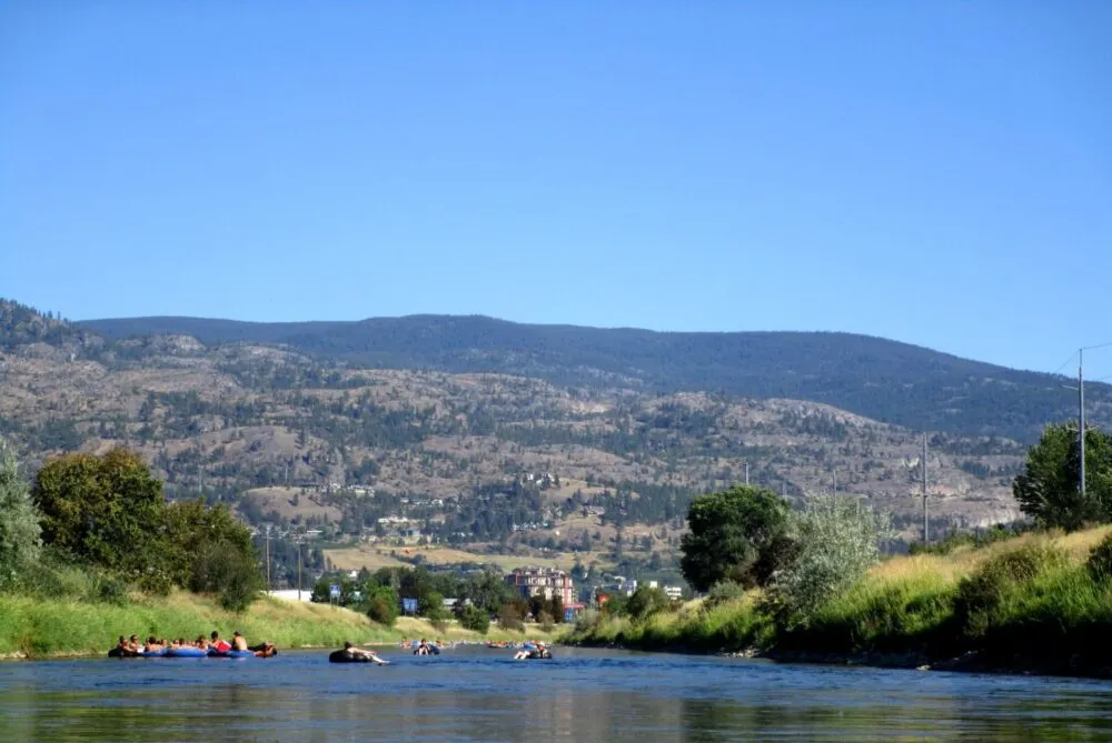 River level view of people in tubes floating down the Penticton Channel, with sunny skies above