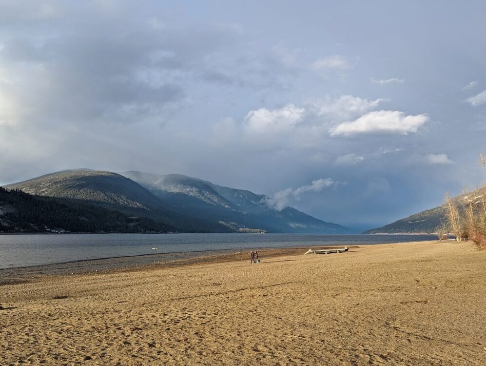 Golden hour on sandy Canoe Beach with people strolling across. Forested mountains on the other side of the lake with stormy weather in distance