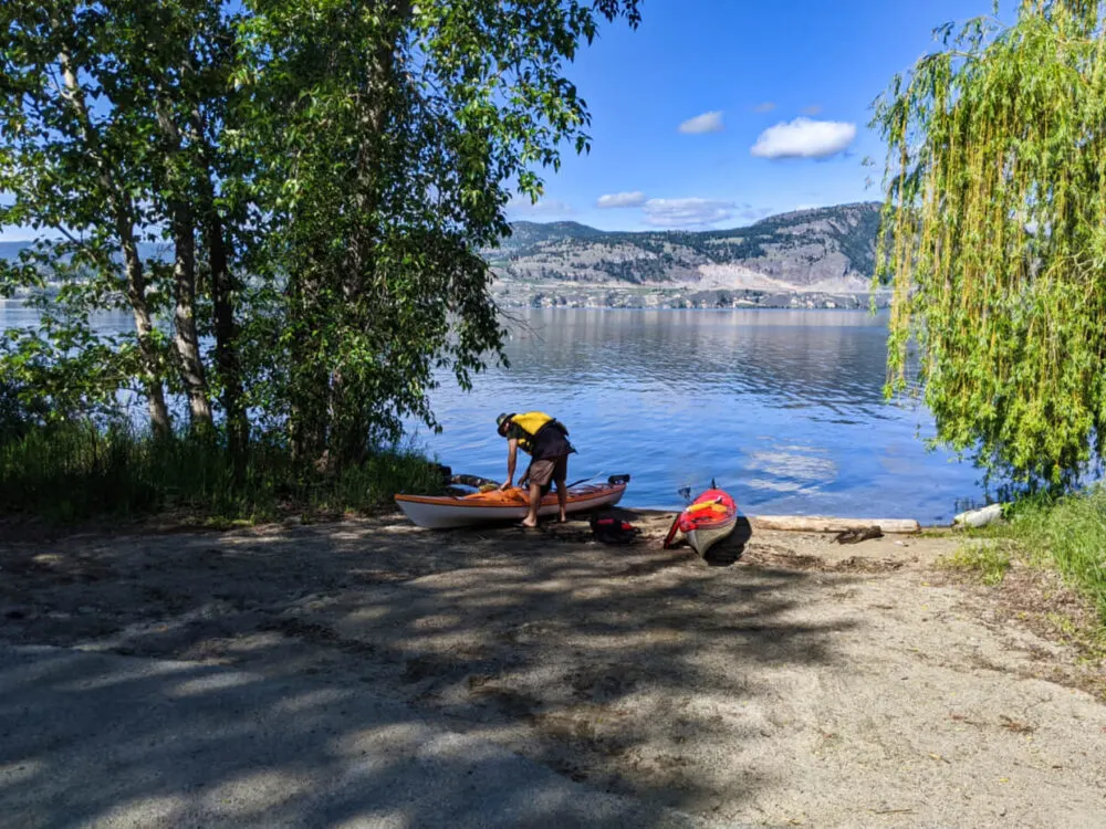 Orange and red kayaks sat on shore next to the lake at Indian Rock boat launch. JR is standing over orange kayak and putting items in storage compartments