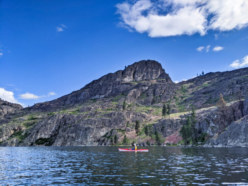 JR is in a red kayak, paddling past the shoreline of Okanagan Mountain Park, which features rugged and rocky scenery with few trees. The lake's surface is very calm