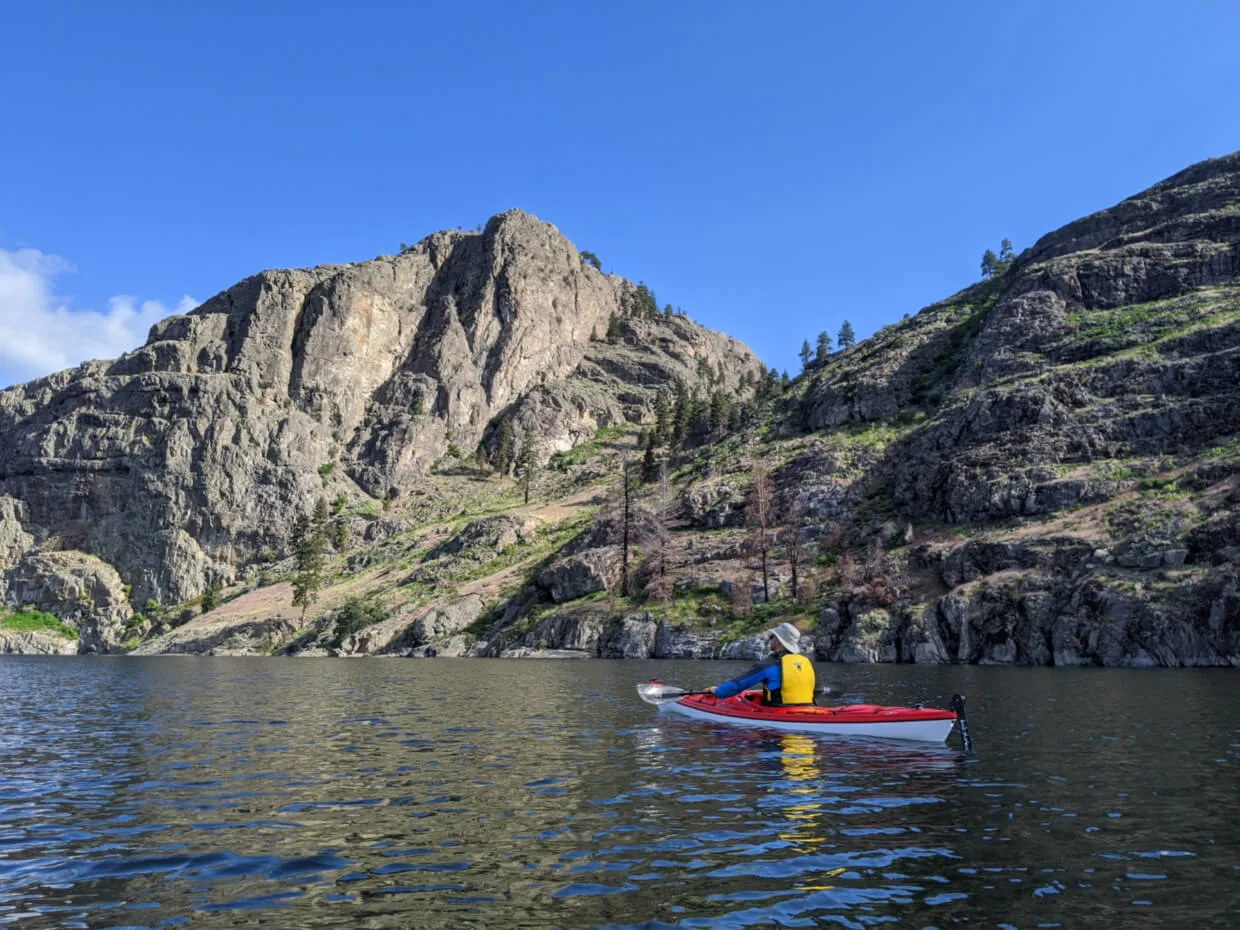 JR sits in red kayak on calm Okanagan Lake, paddling close to the rugged shore of Okanagan Lake