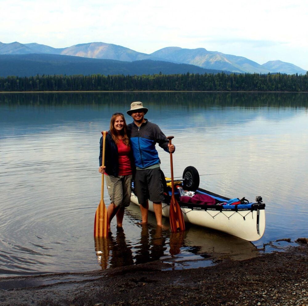 Gemma and JR standing with canoe paddles next to canoe on calm lake