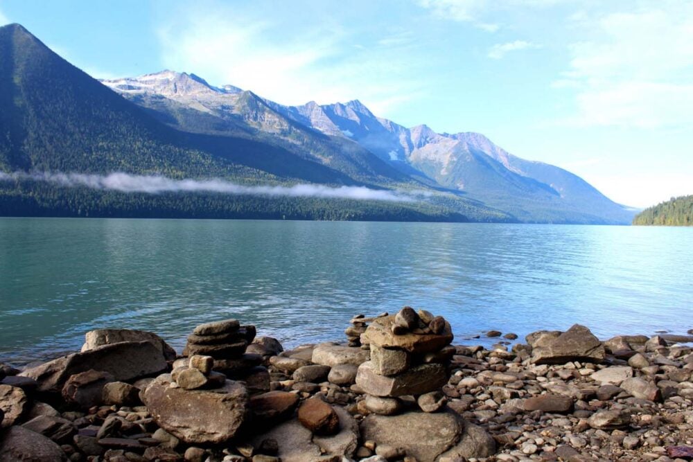 View from shore of pale aquarmine lake bordered by mountains on the Bowron Lake Canoe Circuit, with rock structures on beach in foreground