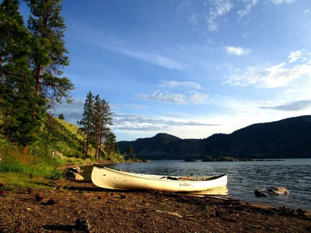 White canoe sitting on shore at sunset, with calm lake and mountainous backdrop