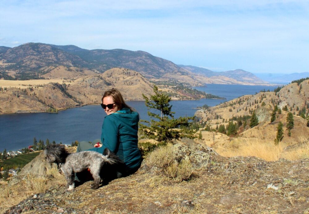 Side view of Gemma and small dog sat on ground in front of scenic lake views at the top of Peach Cliff