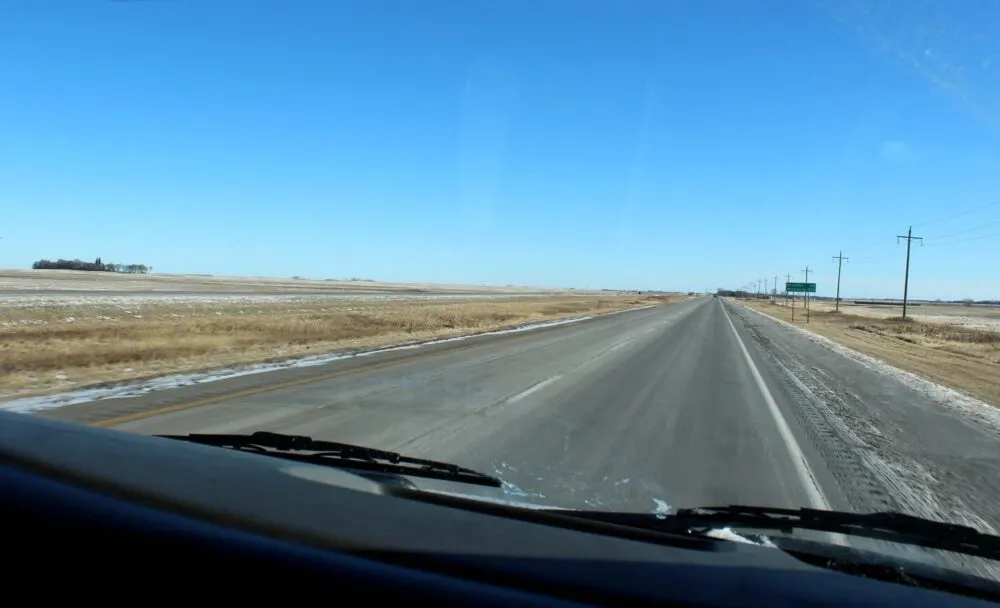 Windshield view of driving through the Canadian prairies with flat brown landscape, small strip of ice on road