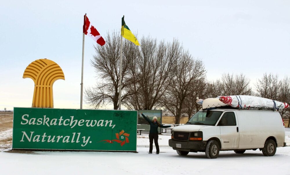 Gemma is standing next to white van at Saskatchewan border in winter, which is marked by a large yellow and green sign