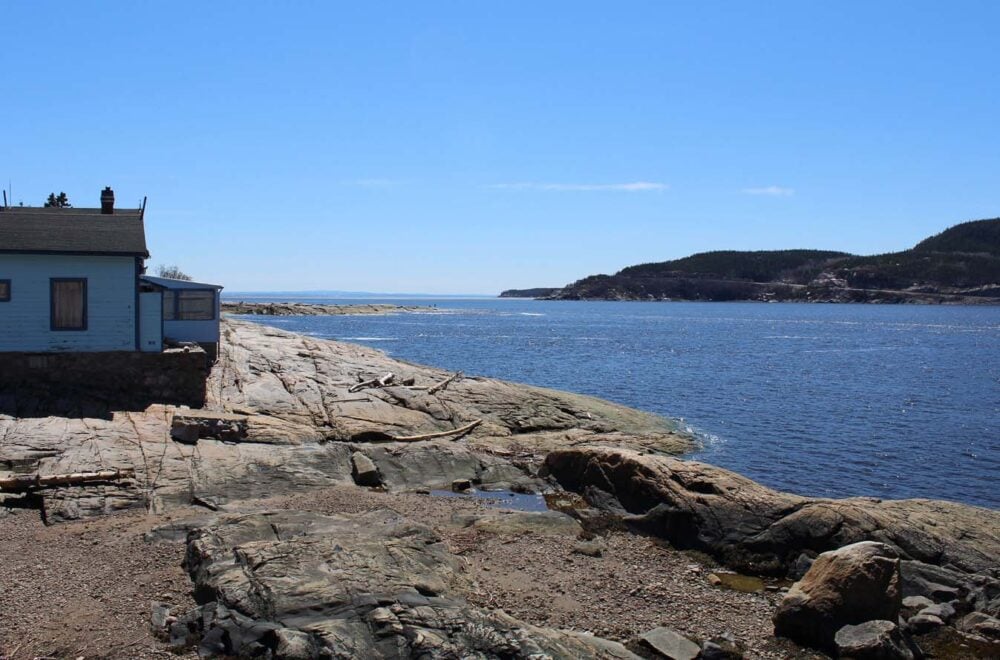 Looking across rocky shoreline with small house on the left, with land visible on other side of river in background