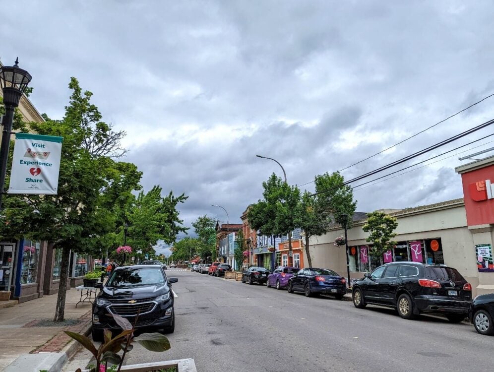 Looking down the main street of Wolfville's downtown area, with cars parked on both sides, with trees above and shops either side