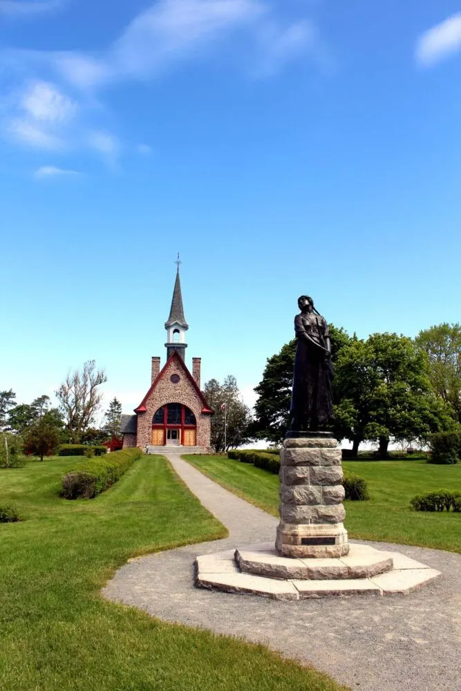 The church at Grand-Pré in background, with high spire. There is a statue of a woman in the foreground (Evangeline)