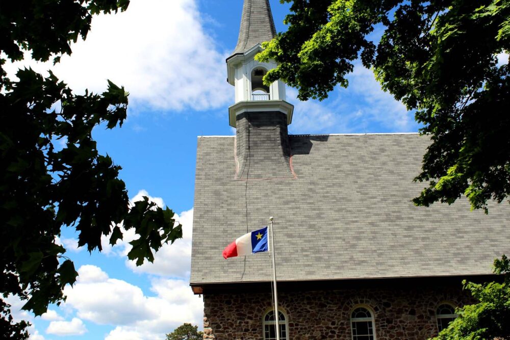 Looking through foliage to the building and spire of the Commemorative Church. There is an Acadian flag (tricolour with yellow star) in the foreground