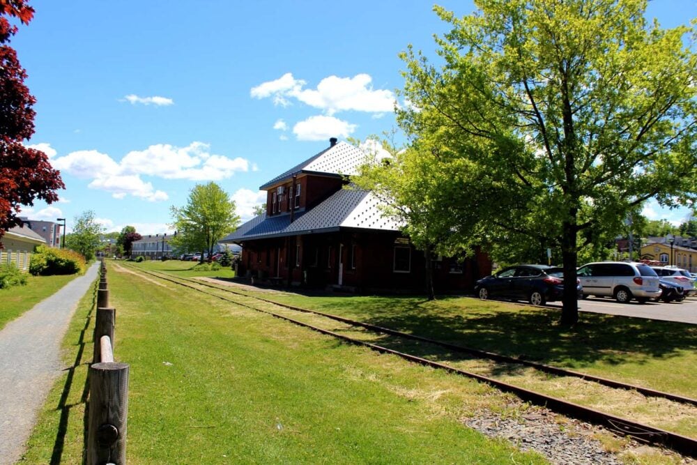 Looking across grassy space towards heritage railway building, now converted into a library. Railway tracks are visible in the grass