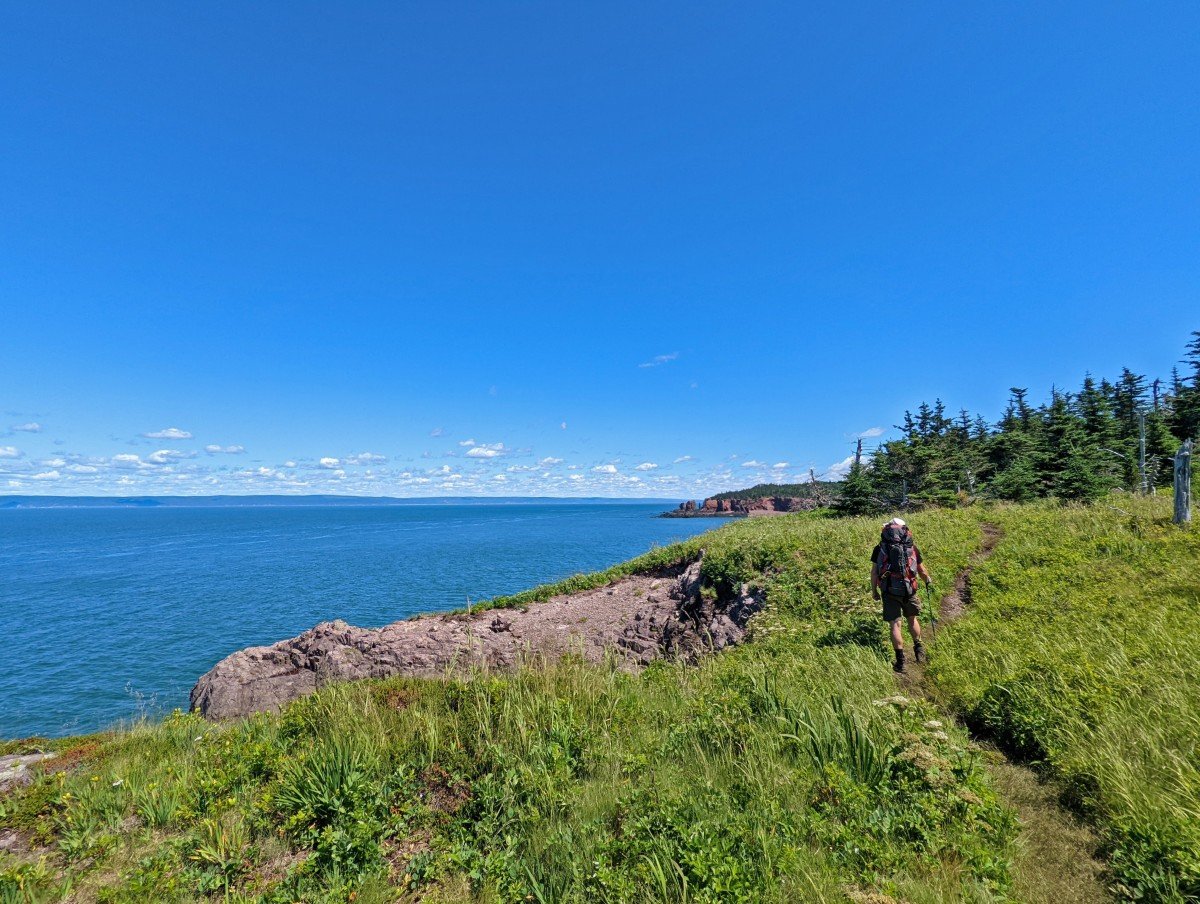 Back view of hiker on open section of the Cape Chignecto Trail, with calm ocean to left, trail head and cliffs visible in distance. Forest on right