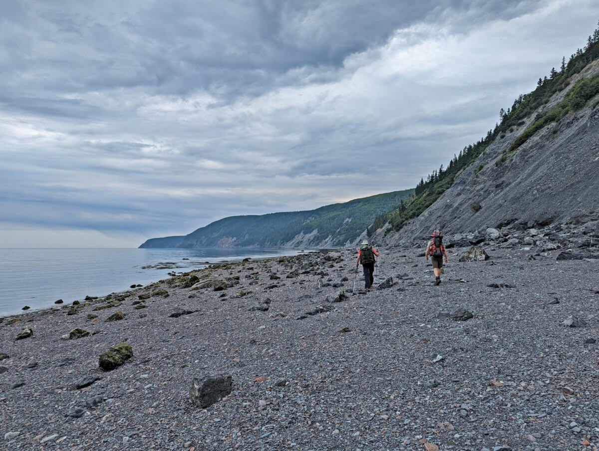 Back view of hikers on Red Rocks Beach, Cape Chignecto Provincial Park, a pebble beach with calm ocean on the left and cliffs on the right