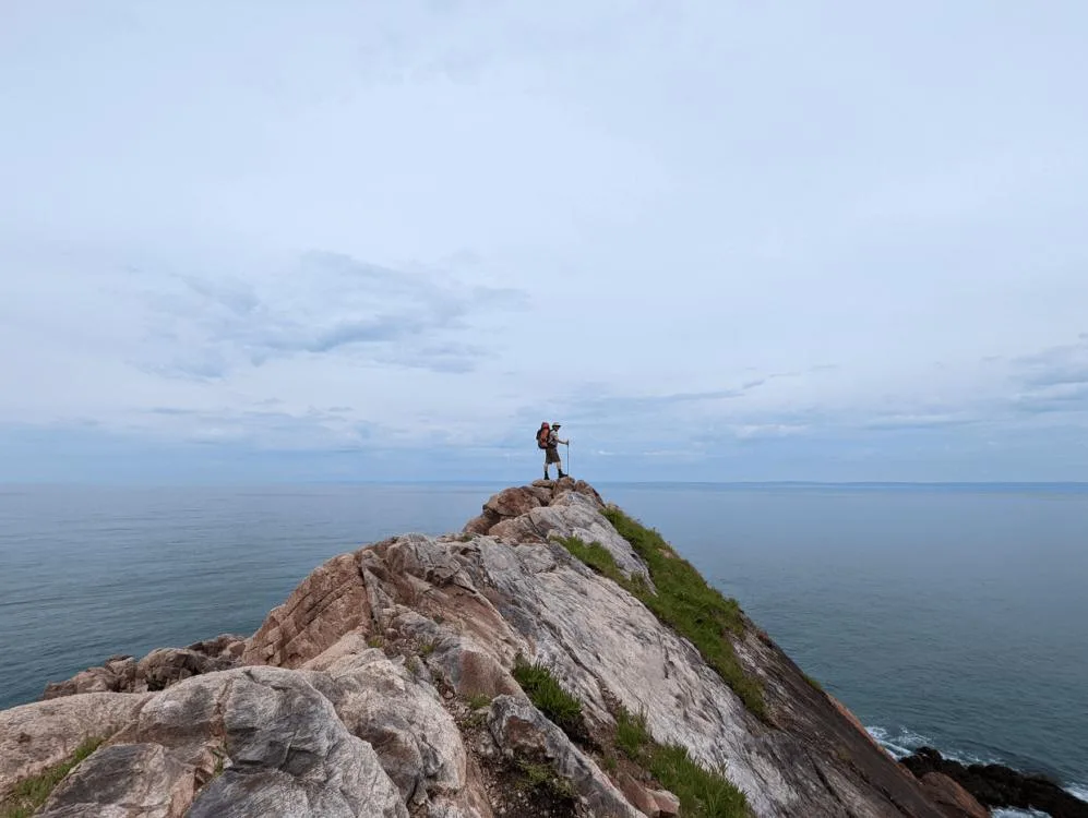 JR standing on sloped rock in front of Bay of Fundy on Cape Chignecto Coastal Trail in Nova Scotia. The ocean is calm