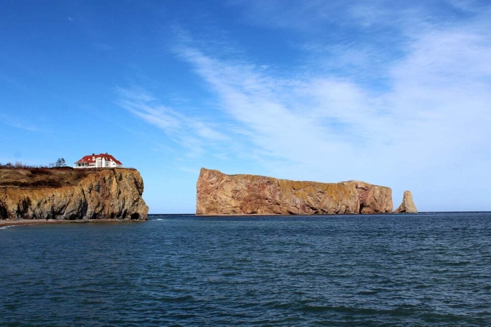 Shark fin shaped Percé Rock separated from mainland by ocean, with white and red house on nearby cliff