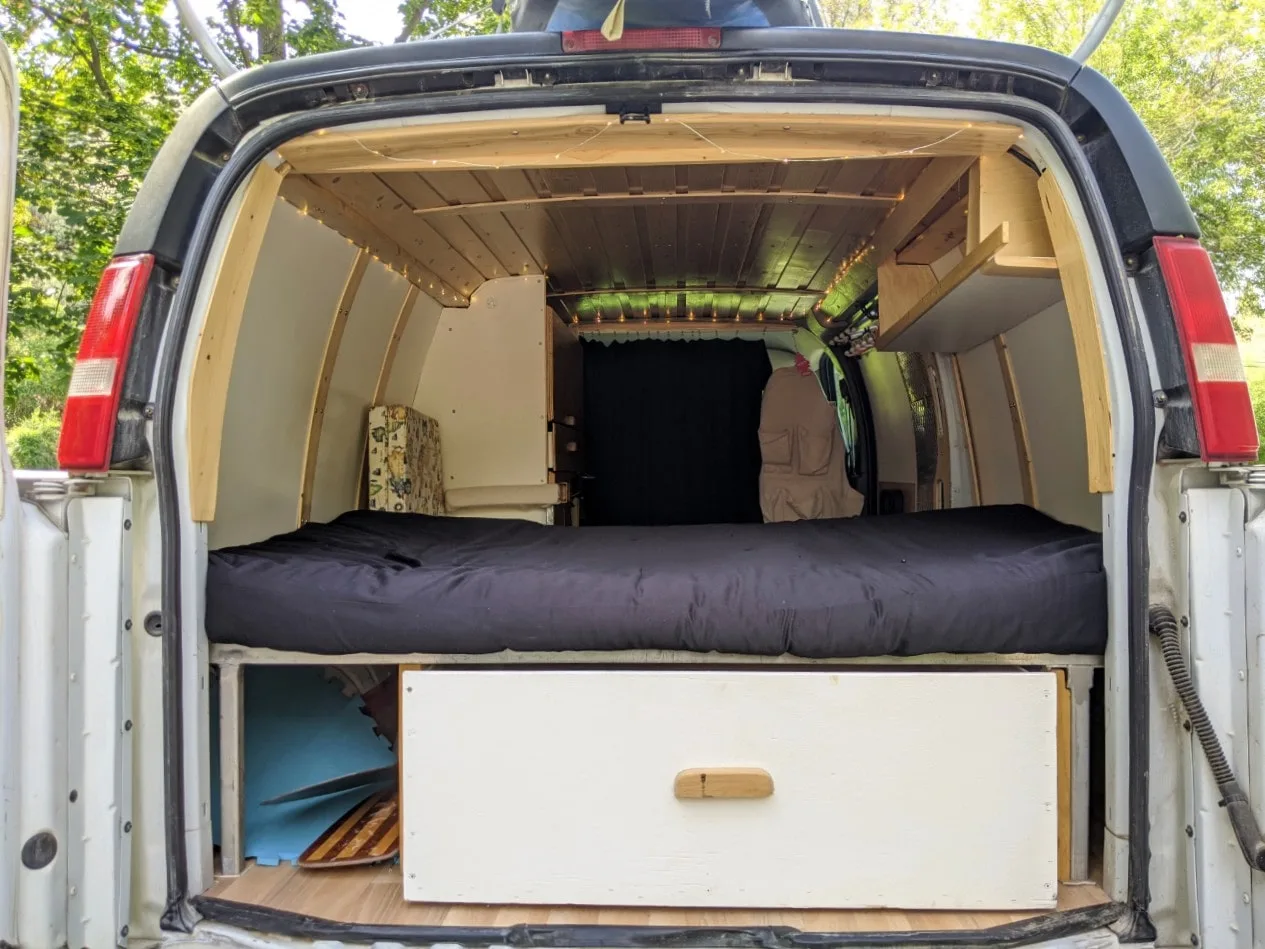 Back view looking into DIY GMC Van conversion, with blue mattress, large white drawer and wooden ceiling