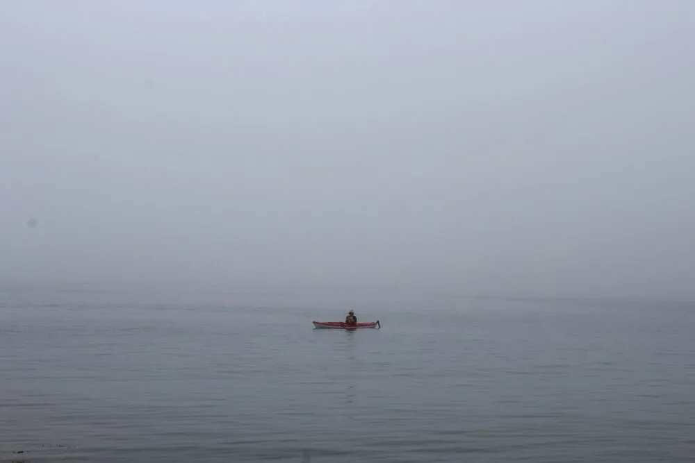 A red kayak floats on a calm ocean surrounded by fog