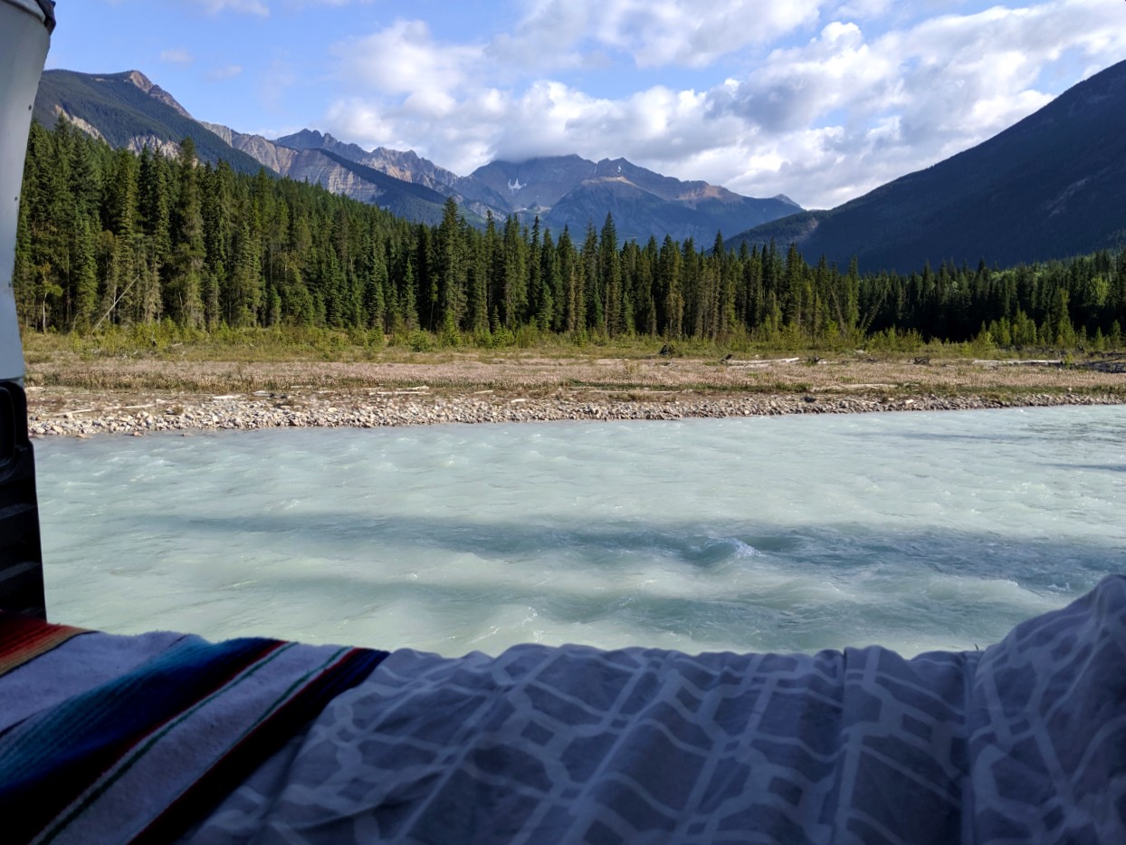 Looking out to a pale river backdropped by mountains from a van with bedsheets and blanket