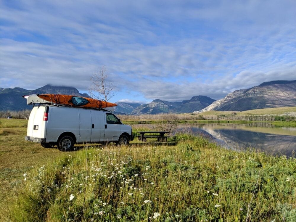 White van parked by picnic table next to reflective lake and mountains