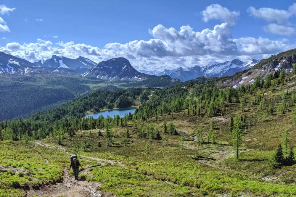 Gemma is hiking up a hill on a dirt trail, heading towards the camera, with lake in background and a number of impressive peaks