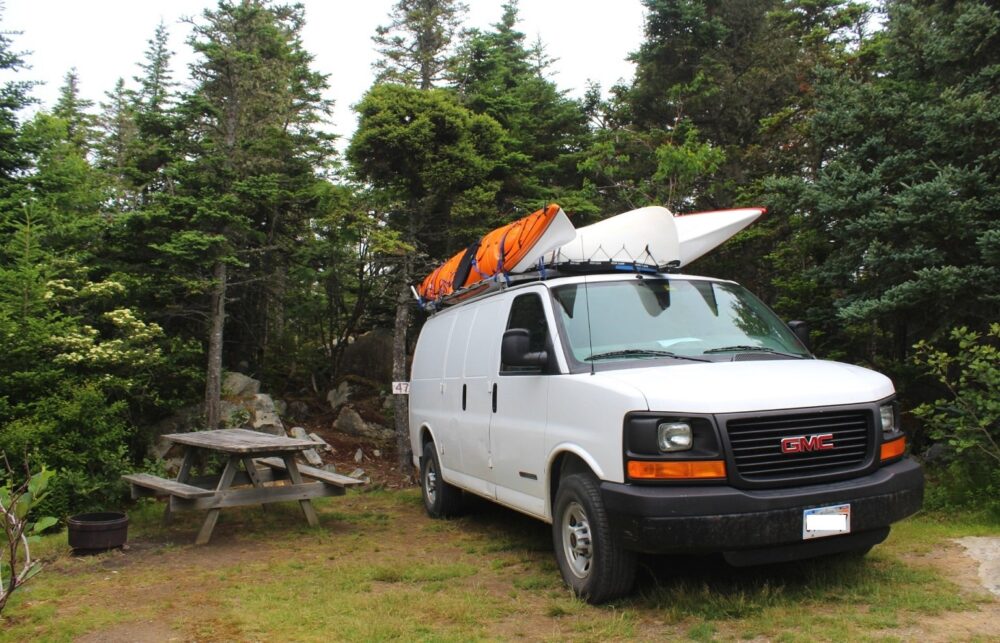 White van parked next to picnic table and firepit, surrounded by forest