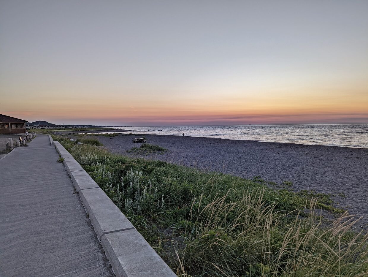 Sunset views on Sainte-Anne-Des-Monts waterfront, with boardwalk on left and sandy/rocky beach on right, separated by dune foliage