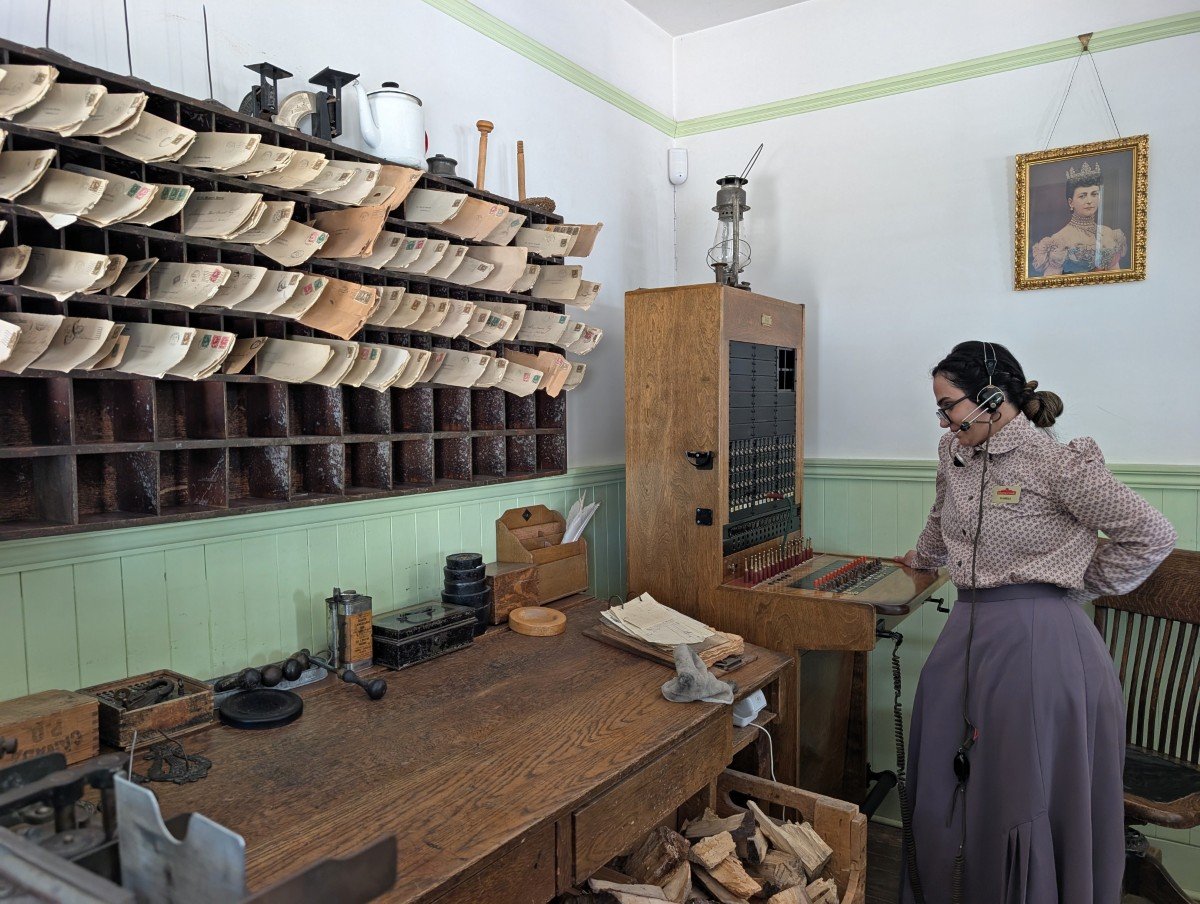 Side view of 19th century dressed actor in Heritage Park's Post Office, listening to phone system. There is a large desk on the left with postal shelves above