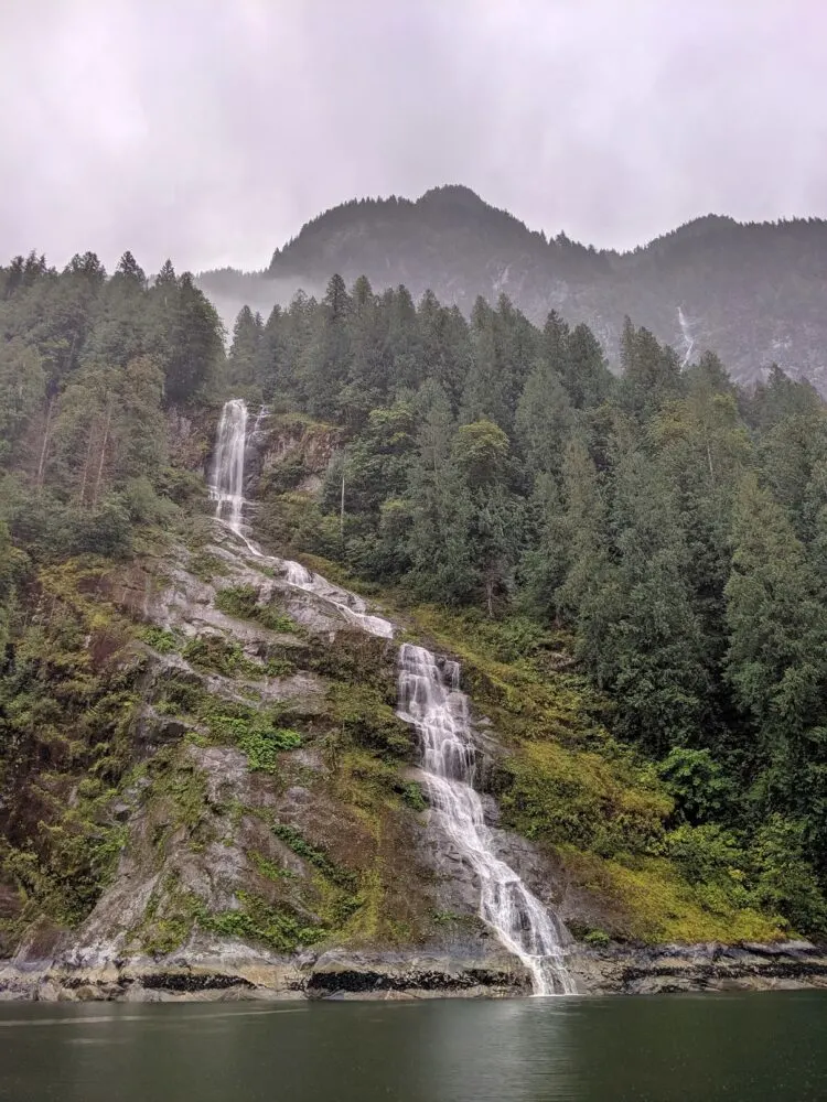 A waterfall cascades down granite fjord walls, surrounded by forest and mist