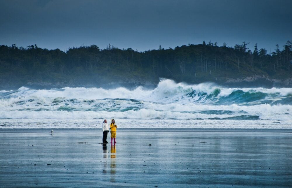 Two people stand on the beach and watch huge storm season waves