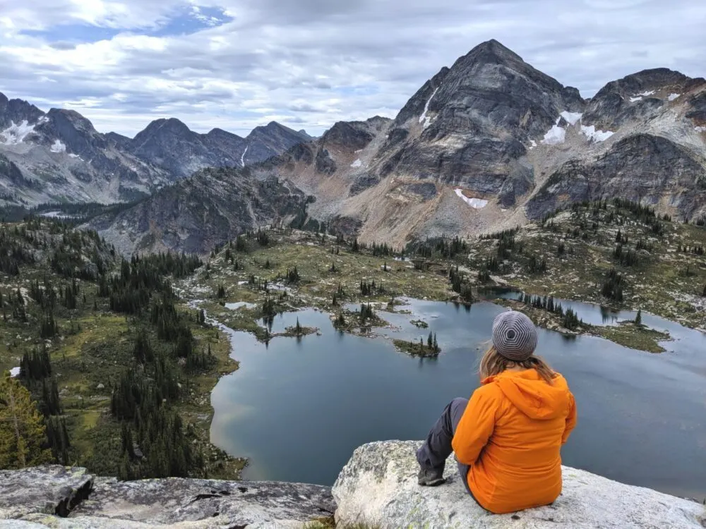 Back view of Gemma sat on rocky wearing orange jacket looking down to lake below, which is surrounded by mountains