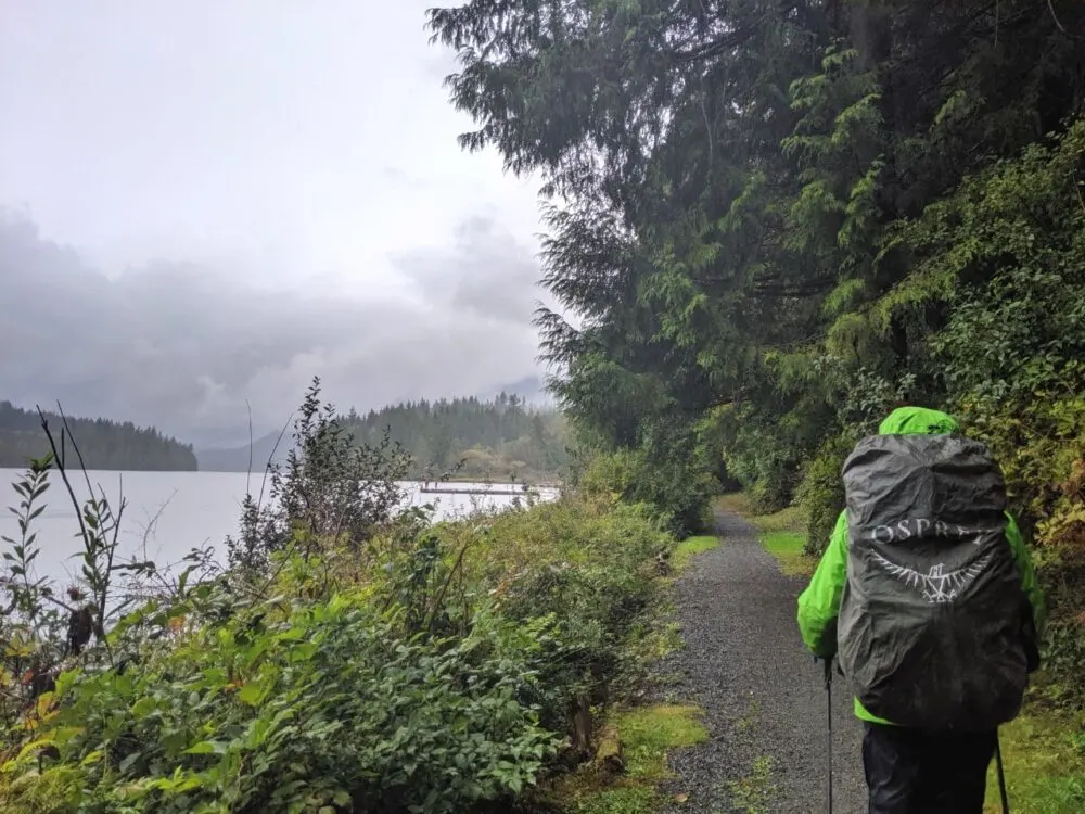 JR hiking next to lake on Sunshine Coast Trail with green rainjacket, hiking poles and an Osprey backpack raincover