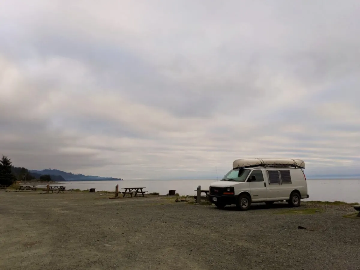 Large gravel campground next to the ocean with picnic tables and fire pits. A white van is parked next to the one of the sites