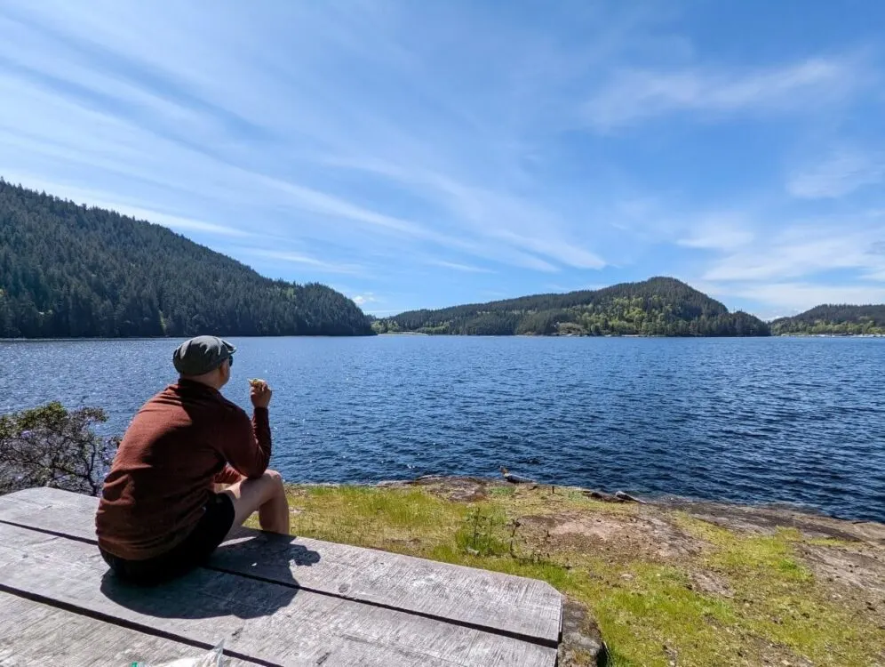 Back view of JR sat down on rock looking across Powell Lake while he eats a sandwich. There are forested hills visible around Powell Lake