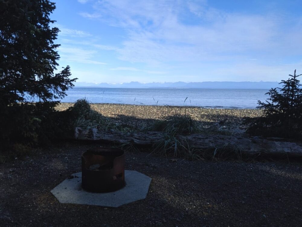 Campfire ring in front of a stony beach, calm ocean and mountains behind