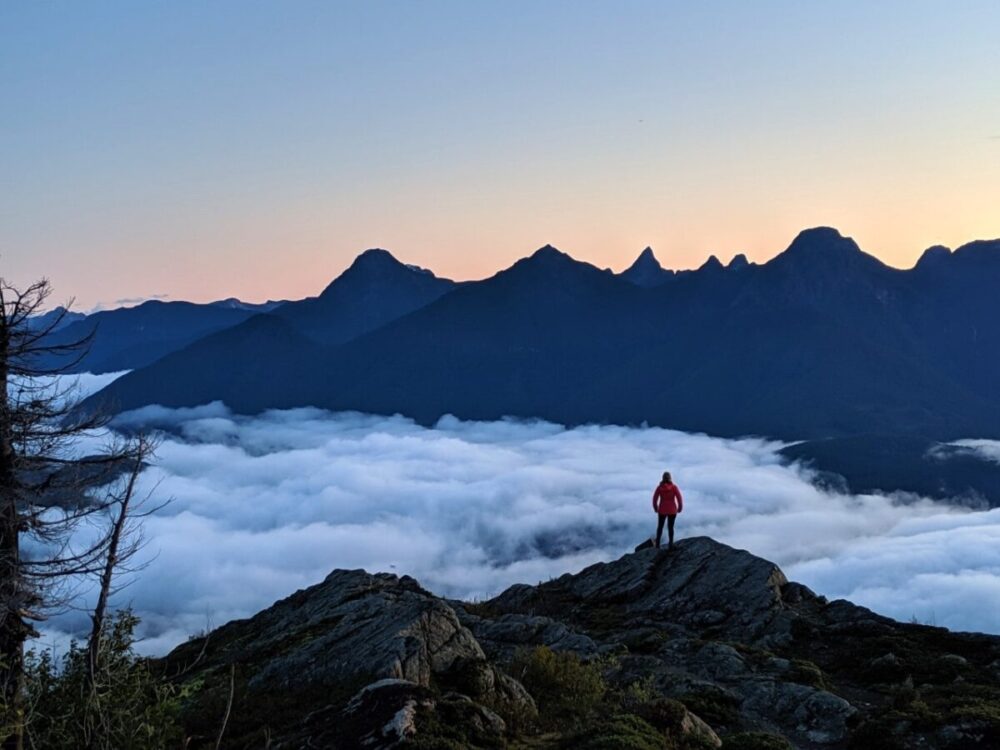 Gemma standing on top of a mountain with sun rising behind peaks, cloud blocking the land below