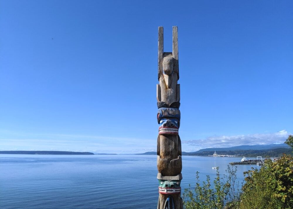 Wooden totem (some painted) in front of coastal scenery in Powell River on the Sunshine Coast. Islands and layers of land are visible in the background