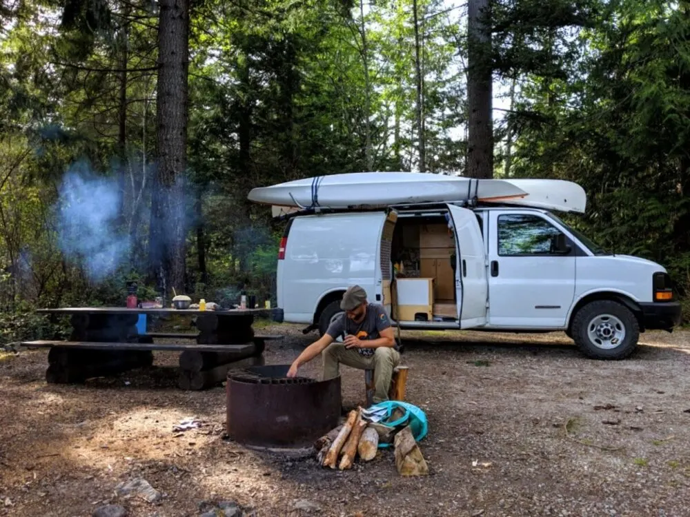 A white van (with one open door) is parked in front of a picnic table and fire pit, where JR is tending a campfire