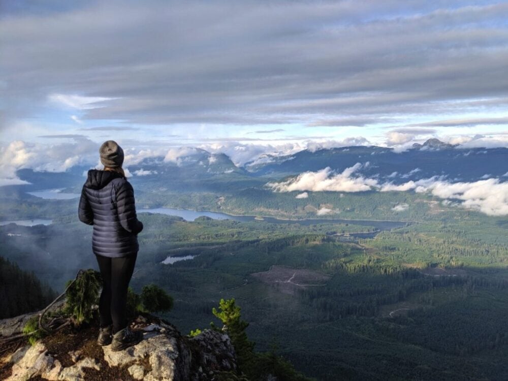 Gemma is standing with back to camera, taking in the views from a cliff looking out over lakes, mountain and forest on a backpacking trip along the Sunshine Coast Trail