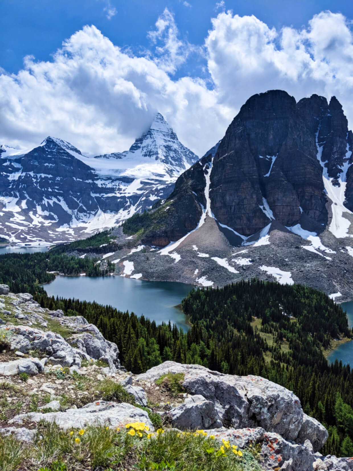 Elevated view looking down towards snowy pyramidal peak in background, with lakes visible in front. Trees surround the lakes