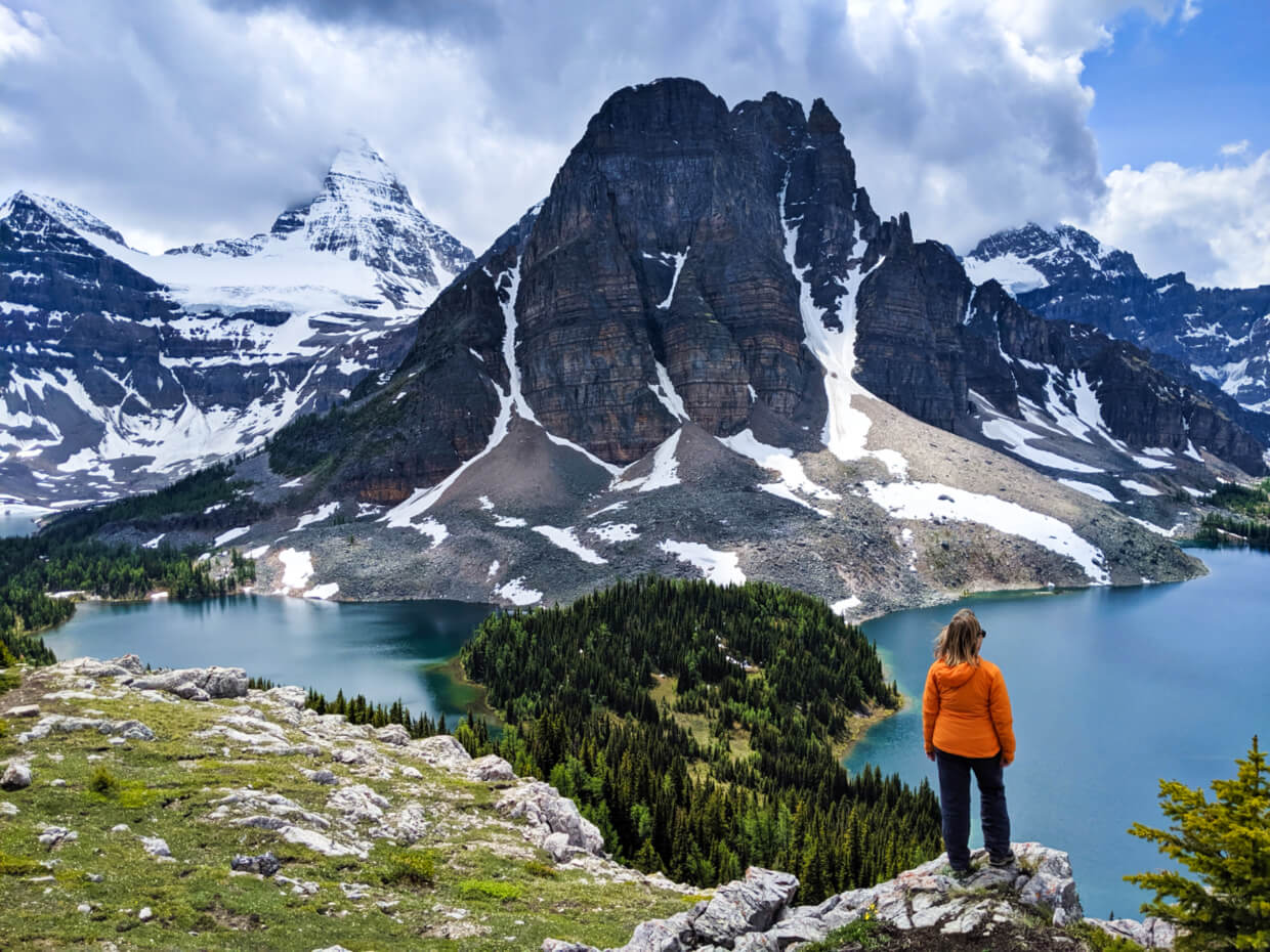 Gemma is standing in an orange jacket in front of a view with a turquoise lake surrounded by mountains