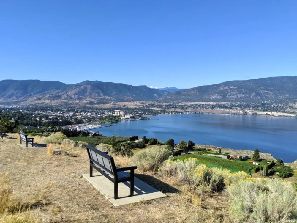 A bench is in the foreground, looking out to Okanagan Lake and the city of Penticton resting on the eastern shore. The city is backdropped by mountains