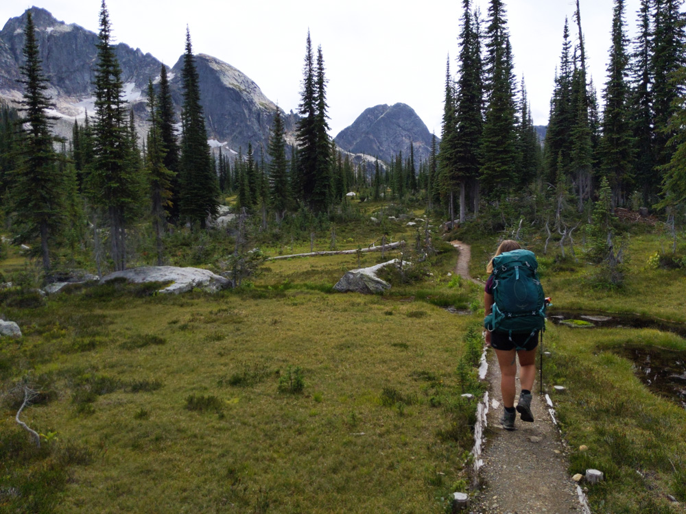 Gemma hikes away from camera on hiking path, carry large backpack. There are mountain peaks in the distance