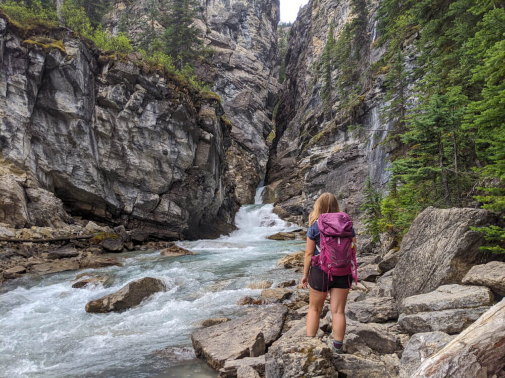 Gemma stands wearing pink backpack with back to camera, standing on rocky surface looking at water rushing through rocky canyon