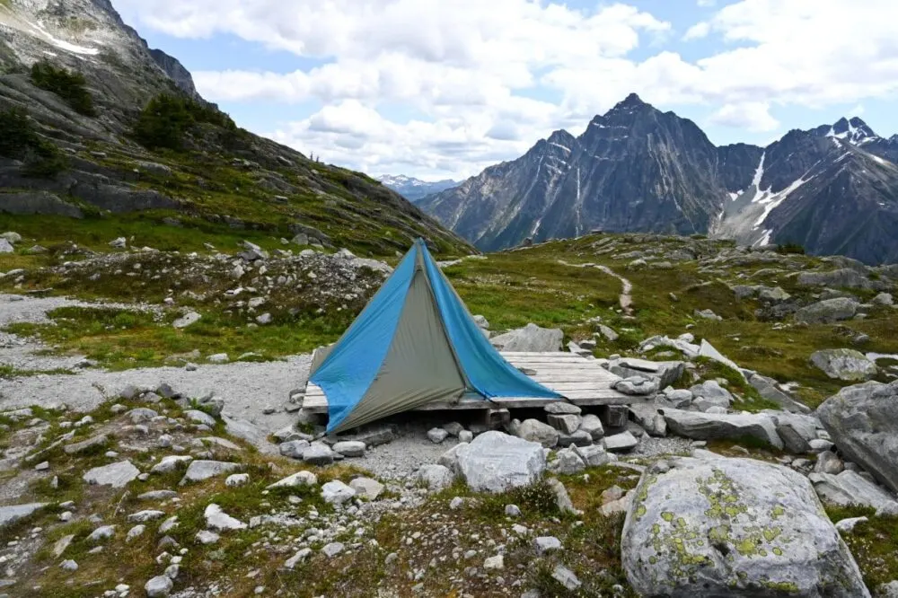 Blue and grey tent on wooden tent pad in alpine area with meadows and mountain backdrop in Glacier National Park, Canada