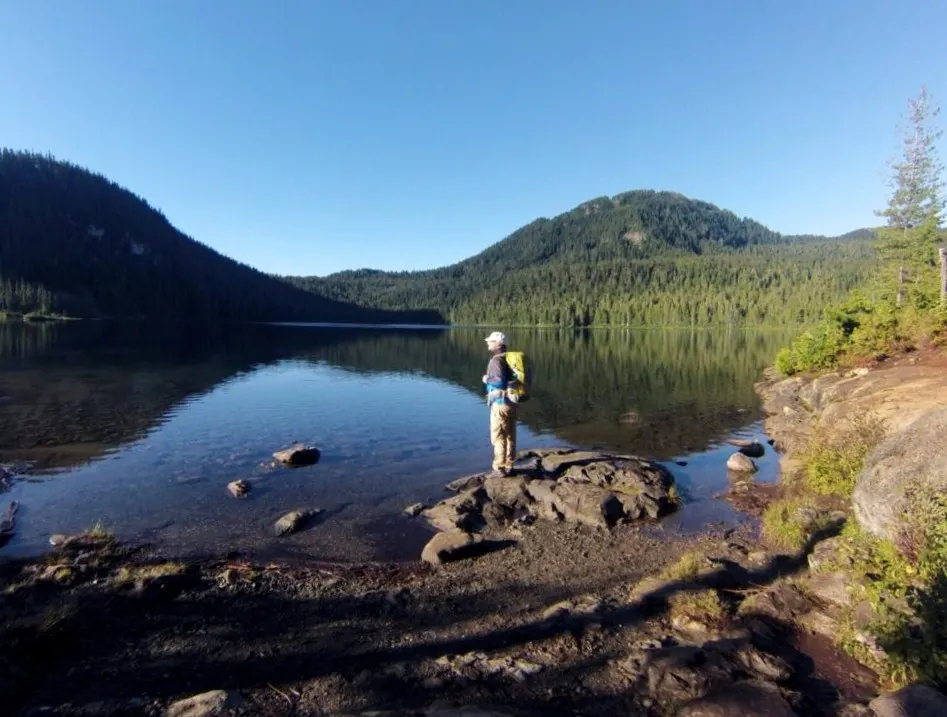 Side profile of hiker standing on rock next to calm lake in Strathcona Provincial Park, with forested hills in the background